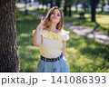 Teen girl fixing windblown hair, standing confidently beside leafy tree in sunlit park during summer afternoon 141086393