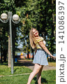 Blonde woman walking through sunny park, wearing yellow top and denim skirt, smiling brightly 141086397