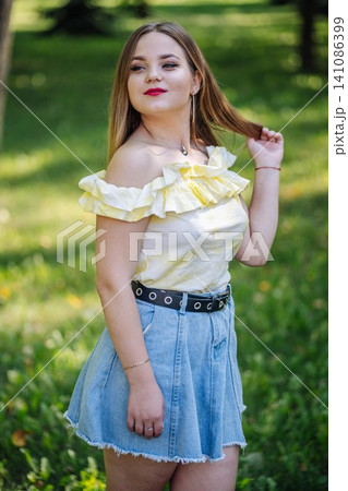 Fashionable young woman wearing a light yellow blouse and a denim skirt, touching her hair in a park, enjoying a sunny summer day Fashionable young woman wearing a light yellow blouse and a denim skirt, touching her hair in a park, enjoying a sunny summer day 141086399