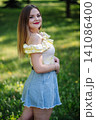 Smiling young woman wearing a light yellow summer dress and denim skirt, posing outdoors in a park 141086400