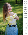 Teenage girl smiling brightly, wearing yellow top and denim skirt, relaxing on grassy park lawn during sunny afternoon 141086402