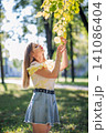 Teenager girl enjoying nature in a park during a sunny autumn day, touching colorful leaves on a branch 141086404