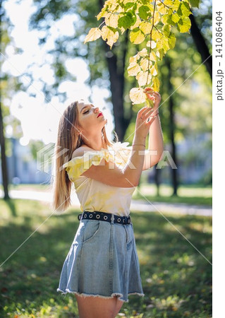 Teenager girl enjoying nature in a park during a sunny autumn day, touching colorful leaves on a branch 141086404