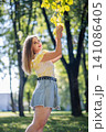 Young woman smiling, wearing trendy outfit, reaching up to colorful autumn foliage during sunny park afternoon 141086405