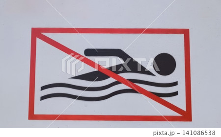 White sign with red border and diagonal line crossing a black silhouette of a swimmer in water, indicating swimming is prohibited. White sign with red border and diagonal line crossing a black silhouette of a swimmer in water, indicating swimming is prohibited. 141086538