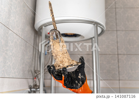 Plumber Holding Removed Water Heater Heating Element Covered With Heavy Mineral Deposits, Showing Hard Water Damage And Maintenance Service Process Plumber Holding Removed Water Heater Heating Element Covered With Heavy Mineral Deposits, Showing Hard Water Damage And Maintenance Service Process 141087116