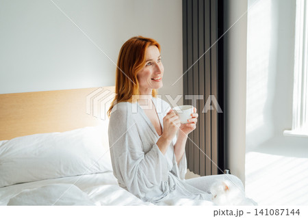 Relaxed and calm middle aged woman with cup of coffee relaxing in bed in morning sunlight. Hot drink for wake up and energy boost. Well being morning routine and daydreaming mood. Good morning habits 141087144