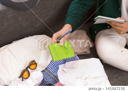 man is packing luggage for a summer vacation. They check a list for travel preparation and place a green passport into the suitcase. man is packing luggage for a summer vacation. They check a list for travel preparation and place a green passport into the suitcase. 141087238