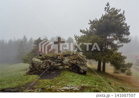 A stone Christian cross stands on a moss-covered rock in a misty, foggy mountain A stone Christian cross stands on a moss-covered rock in a misty, foggy mountain 141087507