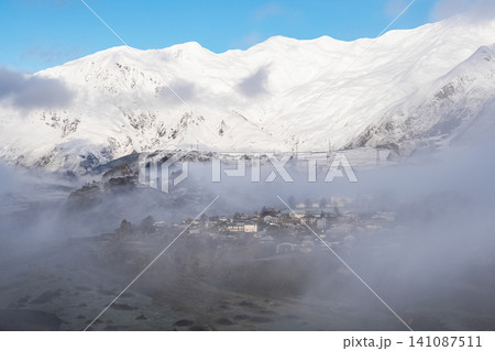 Scenic view of a mountain village surrounded by heavy morning fog and snow-covered peaks, Georgia Scenic view of a mountain village surrounded by heavy morning fog and snow-covered peaks, Georgia 141087511