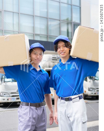 屋外でダンボールを運ぶ制服姿の若い配送員 屋外でダンボールを運ぶ制服姿の若い配送員 141087835