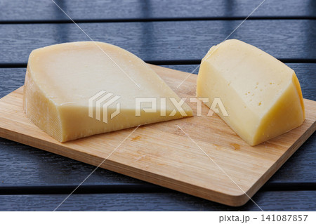 Closeup of two pieces of pale yellow semi hard cheese on a wooden board on a dark table. 141087857