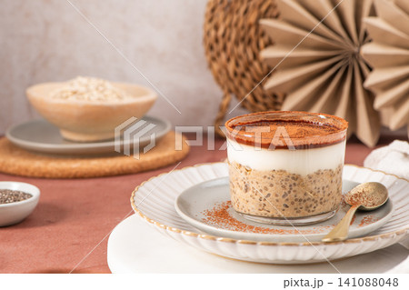 Creamy tiramisu overnight oats and chia pudding topped with a layer of yogurt and dusted with cocoa powder, served in a glass on a white and gray plate with a golden spoon 141088048