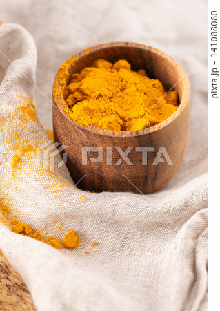 Ground turmeric spice fills a small wooden bowl resting on a textured linen cloth with scattered yellow powder highlights for a natural kitchen aesthetic Ground turmeric spice fills a small wooden bowl resting on a textured linen cloth with scattered yellow powder highlights for a natural kitchen aesthetic 141088080