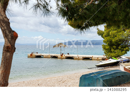 Pebble beachwith a small pier holding boats and a jet ski beside the Adriatic Sea framed by pine branches in Makarska, Croatia. 141088295
