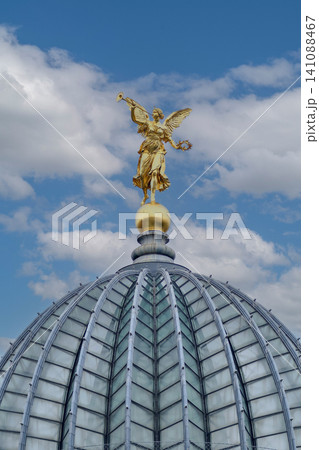 Golden winged angel statue with trumpet standing on top of modern glass building dome against blue sky Golden winged angel statue with trumpet standing on top of modern glass building dome against blue sky 141088467