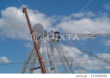 Industrial mobile crane assembling a giant white steel Ferris wheel structure against blue sky with clouds Industrial mobile crane assembling a giant white steel Ferris wheel structure against blue sky with clouds 141088468