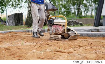 Worker in work boots stands beside vibrating plate compactor on sandy soil near concrete foundation edge, with stacked paving blocks, trees, and outdoor building site background Worker in work boots stands beside vibrating plate compactor on sandy soil near concrete foundation edge, with stacked paving blocks, trees, and outdoor building site background 141089971