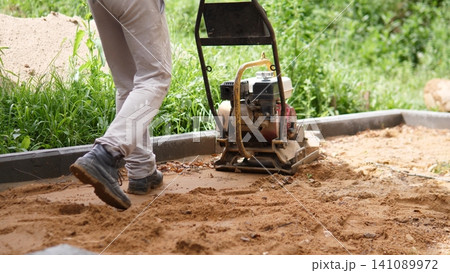 Worker legs beside plate compactor on sandy ground along concrete edging, with lush green vegetation in background, showing site preparation, soil compaction, and groundwork for 141089972