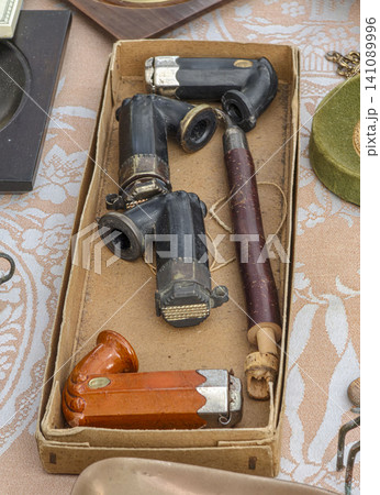 Various antique smoking pipes and vintage pipe parts are displayed in a cardboard box at a flea market. Visitors can browse and purchase unique items. Various antique smoking pipes and vintage pipe parts are displayed in a cardboard box at a flea market. Visitors can browse and purchase unique items. 141089996