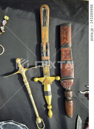 A traditional North African Tuareg dagger lies next to its engraved leather sheath on a black background. The dagger features a detailed brass handle, highlighting its craftsmanship and design. A traditional North African Tuareg dagger lies next to its engraved leather sheath on a black background. The dagger features a detailed brass handle, highlighting its craftsmanship and design. 141089998