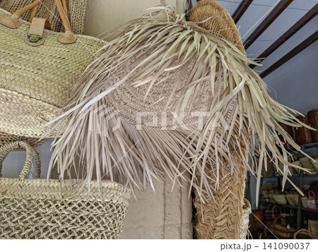 A straw sun hat hangs with frayed edges amid woven wicker baskets in a rustic shop. The scene shows various handmade items on display for sale. A straw sun hat hangs with frayed edges amid woven wicker baskets in a rustic shop. The scene shows various handmade items on display for sale. 141090037