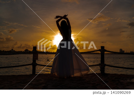 Woman in flowing dress at sunset by ocean Woman in flowing dress at sunset by ocean 141090269