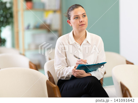 Focused girl student sitting in conference room and writing notes with folder at seminar Focused girl student sitting in conference room and writing notes with folder at seminar 141091642