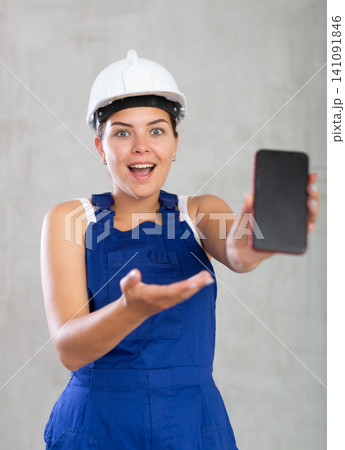 Smiling female worker in work clothes and hardhat demonstrating screen of her phone Smiling female worker in work clothes and hardhat demonstrating screen of her phone 141091846