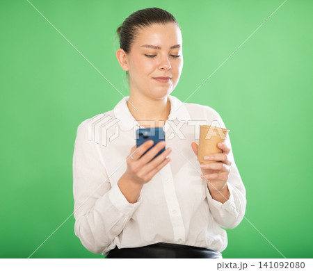 Woman drinking a hot drink while scrolling social networks on a green background Woman drinking a hot drink while scrolling social networks on a green background 141092080