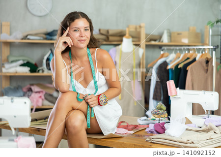 Carefree young female tailor sitting on workbench in workshop Carefree young female tailor sitting on workbench in workshop 141092152