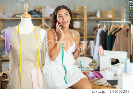 Young clothing designer sitting on table, chatting on smartphone in workshop Young clothing designer sitting on table, chatting on smartphone in workshop 141092222