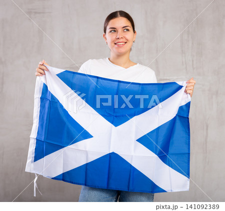 Joyous young woman holding Scotland flag against unicoloured background Joyous young woman holding Scotland flag against unicoloured background 141092389