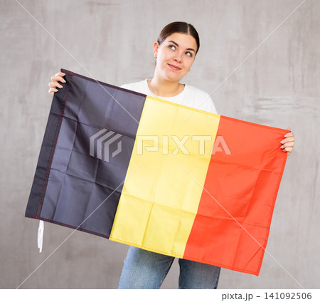 Glad young woman posing and smiling, standing with flag of Belgium against gray wall Glad young woman posing and smiling, standing with flag of Belgium against gray wall 141092506