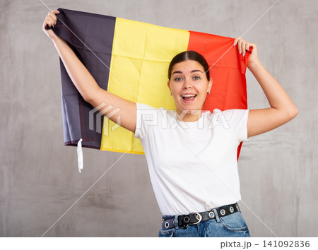 Glad young woman posing and smiling, standing with flag of Belgium against gray wall Glad young woman posing and smiling, standing with flag of Belgium against gray wall 141092836