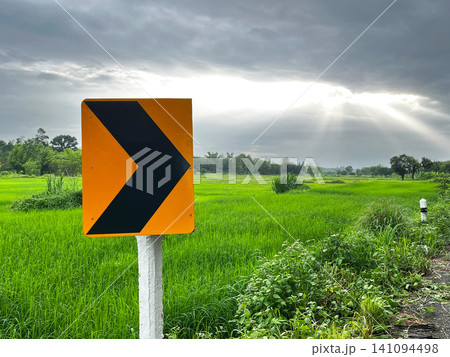 Civil engineering photograph showcasing a yellow chevron alignment sign on a concrete post beside a rural road curve, overlooking green rice paddy fields Civil engineering photograph showcasing a yellow chevron alignment sign on a concrete post beside a rural road curve, overlooking green rice paddy fields 141094498