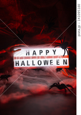 Dark still life featuring illuminated lightbox with HAPPY HALLOWEEN message holiday arrangement. Realistic skeleton arm reaches toward display, while decorative spider web creates spooky Dark still life featuring illuminated lightbox with HAPPY HALLOWEEN message holiday arrangement. Realistic skeleton arm reaches toward display, while decorative spider web creates spooky 141095180