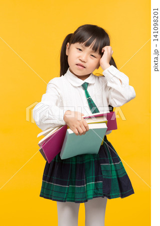 a confused schoolgirl scratching her head while holding textbooks against a yellow background. a confused schoolgirl scratching her head while holding textbooks against a yellow background. 141098201