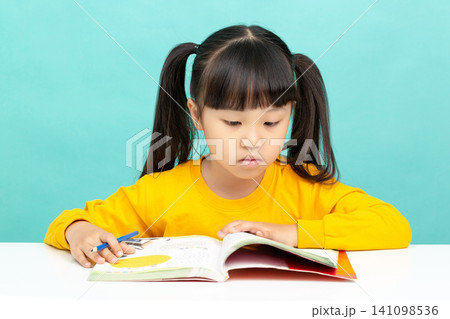 An Asian female student is reading in a blue background. Her expression is serious and focused, suggesting she may have learning difficulties. An Asian female student is reading in a blue background. Her expression is serious and focused, suggesting she may have learning difficulties. 141098536