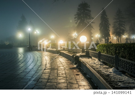 Wooden bench in the snow in a winter night park in the fog. Footpath in a winter city park at night in fog with benches and latterns. Beautiful foggy evening in the Mariinsky Park. Kiev, Ukraine. Wooden bench in the snow in a winter night park in the fog. Footpath in a winter city park at night in fog with benches and latterns. Beautiful foggy evening in the Mariinsky Park. Kiev, Ukraine. 141099141