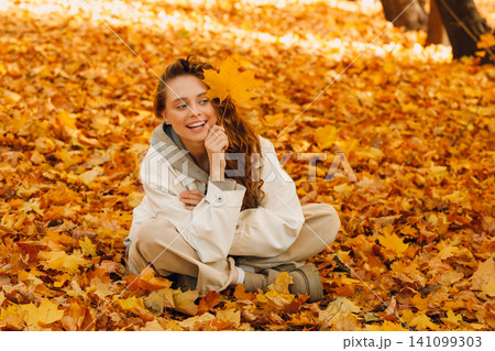 Smiling young woman sits in the autumn foliage on the ground of the forest with the yellow leaves 141099303