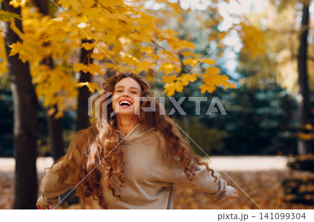 Smiling young woman with curly hairstyle enjoys the autumn weather in the forest with the yellow leaves at sunset Smiling young woman with curly hairstyle enjoys the autumn weather in the forest with the yellow leaves at sunset 141099304