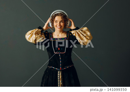 Portrait of a smiling young aristocratic woman with headphones and touching head and dressed in a medieval dress listens music Portrait of a smiling young aristocratic woman with headphones and touching head and dressed in a medieval dress listens music 141099350