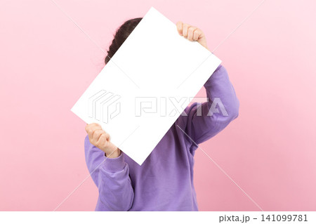 Little girl in purple shirt holding a large blank white horizontal poster with both hands, presenting copy space against a pink wall. 141099781