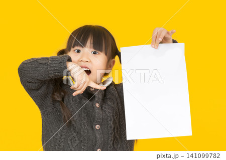 Surprised little girl pointing her finger at a blank white paper sheet with an open mouth expression on a vivid yellow background. 141099782