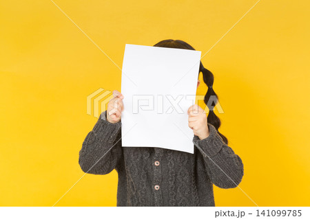 Playful little girl hiding her face behind a blank white sheet of paper, standing against a bright yellow studio background. Playful little girl hiding her face behind a blank white sheet of paper, standing against a bright yellow studio background. 141099785