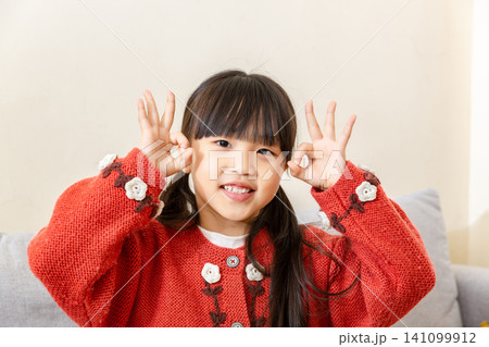 Happy elementary girl standing indoors and making okay signs with both hands near her ears, smiling and looking at camera 141099912