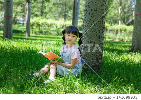 Cute elementary girl with pigtails sitting under a tree trunk in a park and holding an open book, looking at the camera. Cute elementary girl with pigtails sitting under a tree trunk in a park and holding an open book, looking at the camera. 141099935
