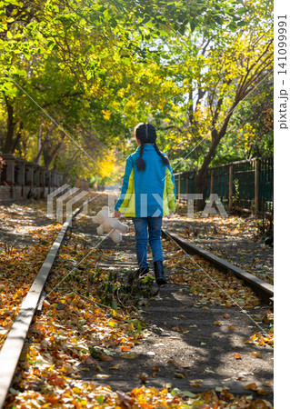 Back view of a child walking down a historic train track carrying a white teddy bear in autumn. 141099991