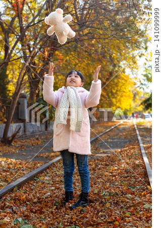 A cheerful young girl throwing a plush white teddy bear up into the air on an autumn railway track. 141099999